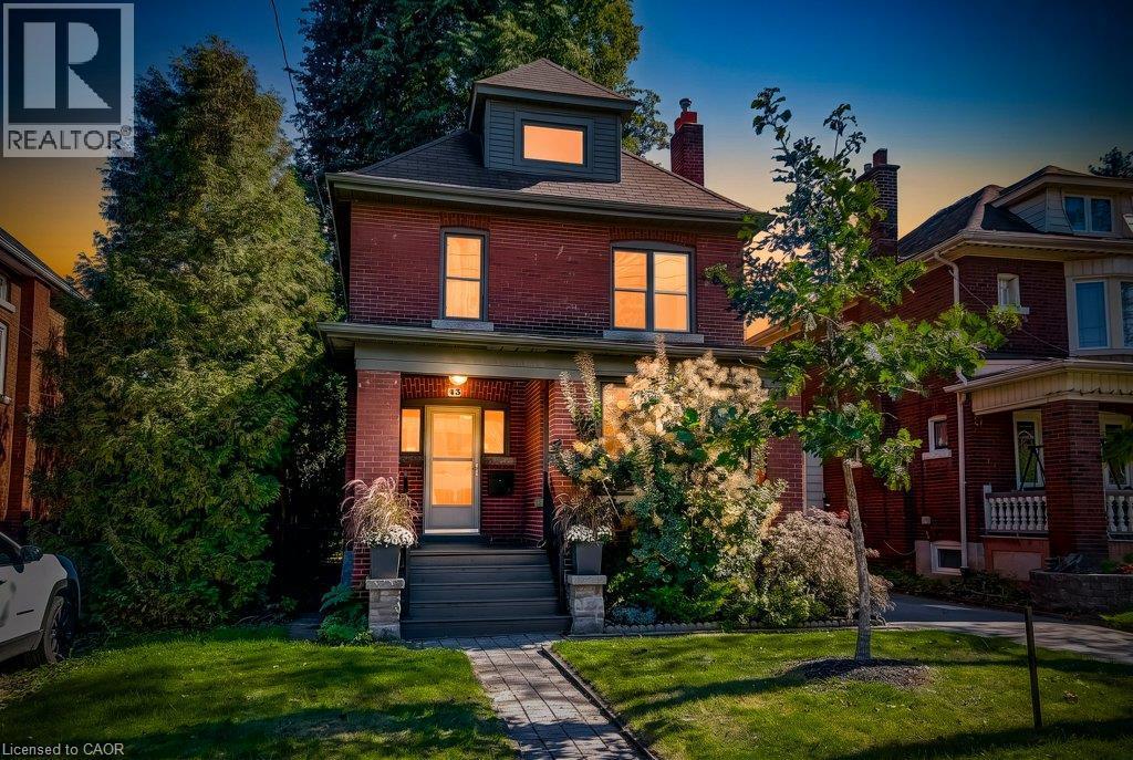 Red brick exterior with a gray-shingled roof, featuring a covered porch with stone-clad pillars, a dormer window, and a paved walkway - 43 Rosslyn Avenue S, Hamilton, ON