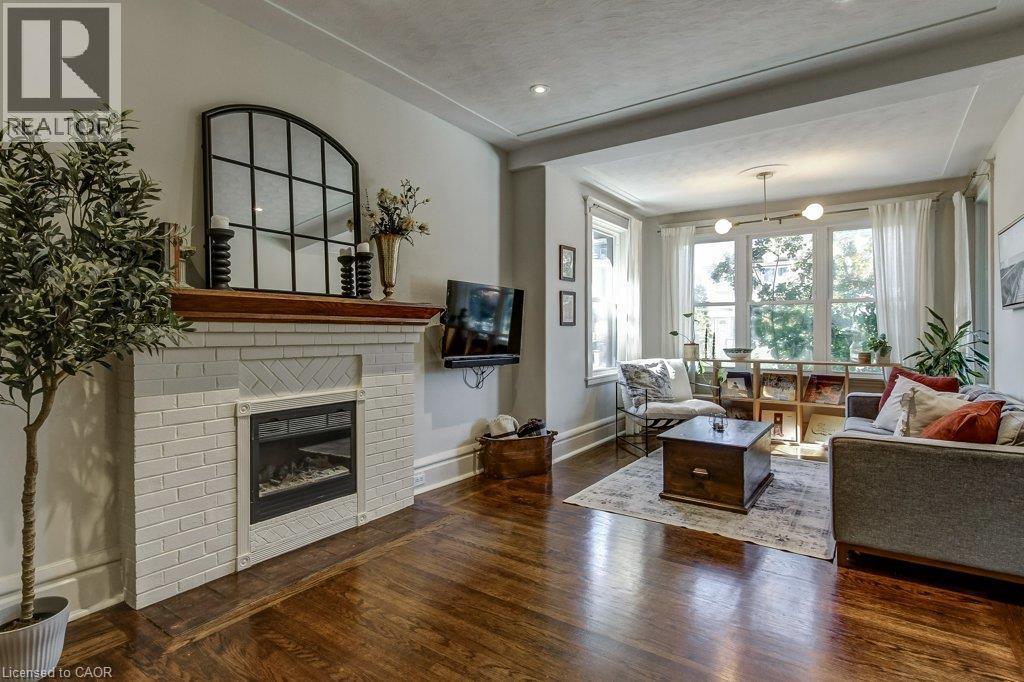 Hardwood flooring, white brick fireplace with mantel, recessed lighting, large windows, and a built-in ceiling light fixture - 43 Rosslyn Avenue S, Hamilton, ON