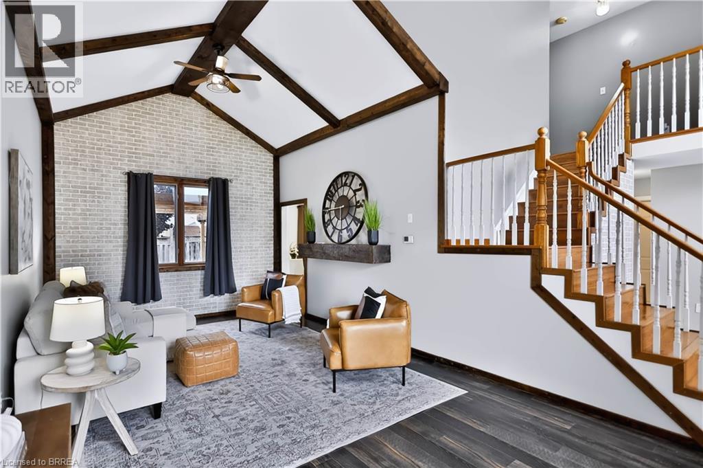 Vaulted ceiling with exposed wood beams, accent brick wall, wood-finish flooring, natural wood staircase with white balusters, and a recessed wall niche - 19 Hyslop Drive, Caledonia, ON