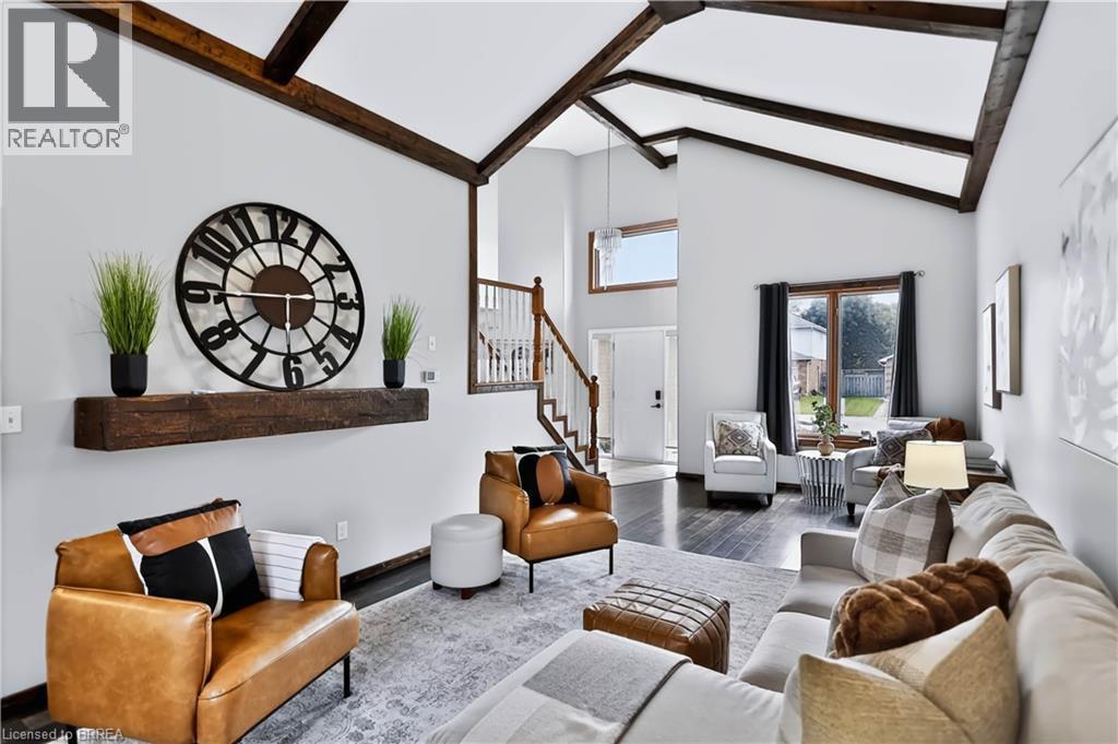 Vaulted ceilings with exposed wood beams, dark wood-finish flooring, staircase with wood handrail and white spindles, transom window, and a large picture window - 19 Hyslop Drive, Caledonia, ON