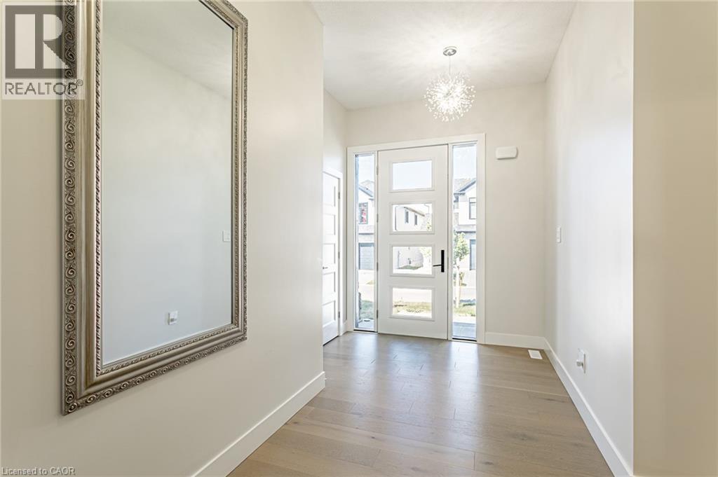 Wood-finish flooring, white walls, modern front door with frosted glass panels, full-height decorative mirror, and a contemporary ceiling light fixture - 7966 Fallon Drive Unit# 18, Lucan Biddulph (Twp), ON - Indoor Photo Showing Other Room