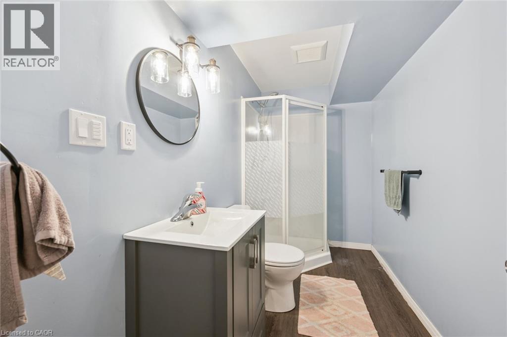 Vanity with integrated sink and chrome faucet, round wall-mounted mirror, three-light sconce, enclosed shower, and wood-finish flooring - 765 Angler Way, Waterloo, ON - Indoor Photo Showing Bathroom