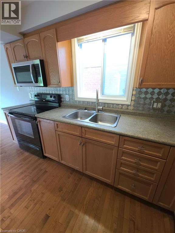 Wood-finish flooring, wood-finish cabinetry, stainless steel double basin sink with a single-handle faucet, integrated microwave, and a decorative backsplash - 998 Mapleridge Street, London, ON - Indoor Photo Showing Kitchen With Double Sink