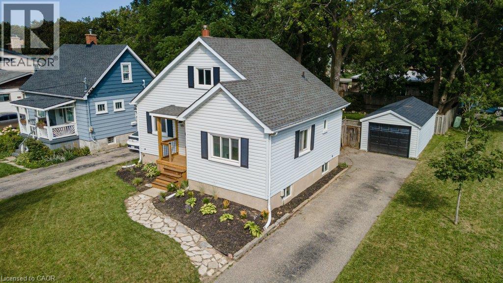 Light-colored siding exterior with dark shutters, covered porch with natural wood railing, stone-finish walkway, detached garage with black garage door, and a paved driveway - 99 Shantz Lane, Kitchener, ON - Outdoor