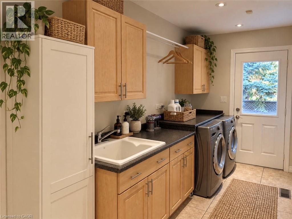 Recessed lighting illuminates the laundry area featuring light wood-finish cabinetry, a utility sink with a chrome faucet, and a dark countertop - 56 Windrush Trail, Kitchener, ON