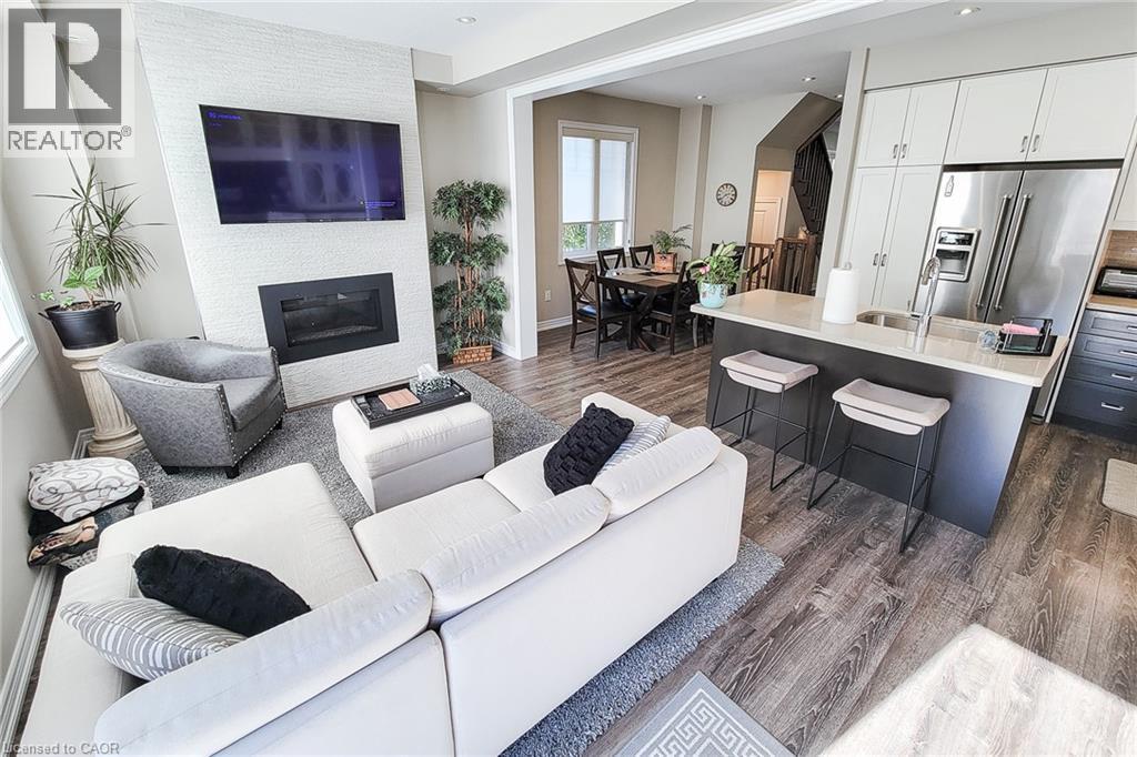 Open-concept living area featuring wood-finish flooring, a contemporary fireplace with a textured surround, and a kitchen island with seating - 65 Waterview Lane, Grimsby, ON