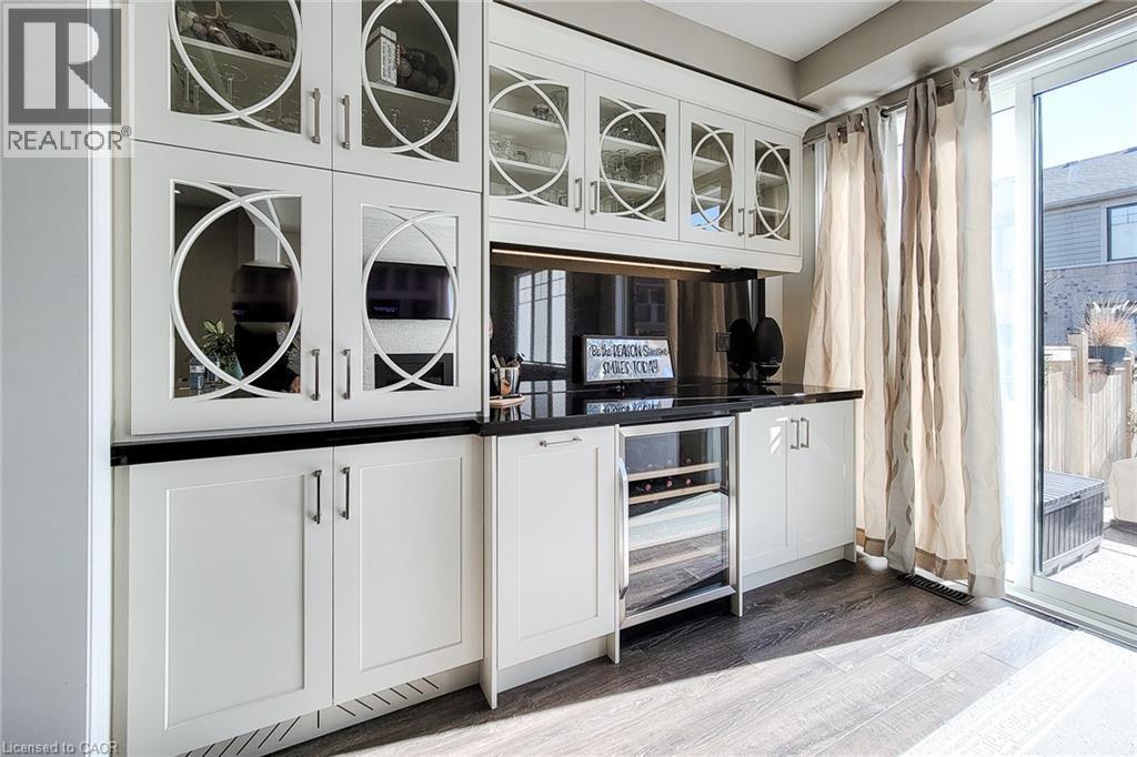 Built-in cabinetry with glass-front uppers and decorative mullions, black countertops, stainless steel backsplash, integrated beverage cooler, and wood-finish flooring - 65 Waterview Lane, Grimsby, ON