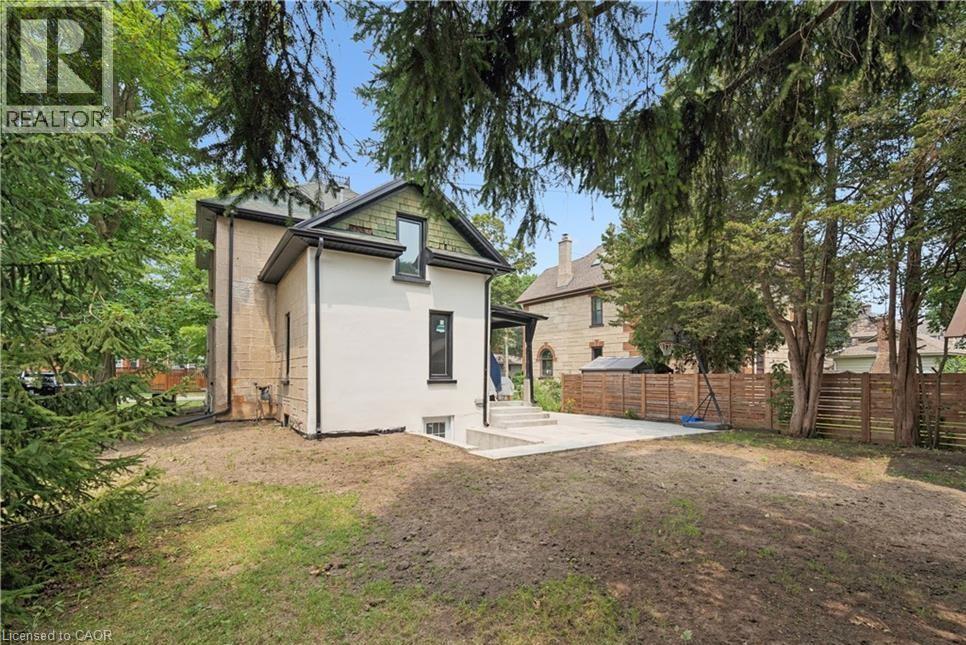 Exterior featuring a neutral stucco finish, dark framed windows, a gabled roofline, a concrete patio, and a wood fence - 245 Lowther Street S, Cambridge, ON