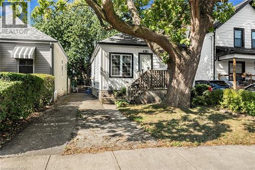White siding exterior with black trim, a mature shade tree, a concrete pathway, a wooden porch with railing, and a striped window awning - 29 Wexford Avenue N, Hamilton, ON 