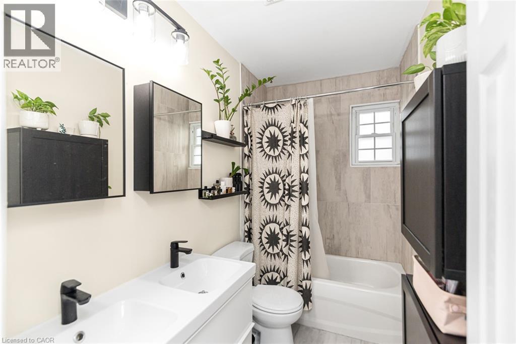 Dual rectangular undermount sinks with matte black faucets, modern vanity with concealed storage, built-in medicine cabinet with mirror, tiled shower surround, and a fixed window - 29 Wexford Avenue N, Hamilton, ON