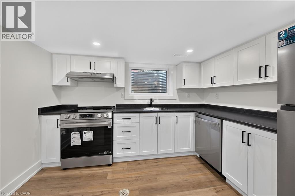 Modern kitchen featuring white shaker cabinetry, dark countertops, wood-finish flooring, stainless steel appliances, and a stainless steel sink with a black faucet - 331 East 17Th Street, Hamilton, ON