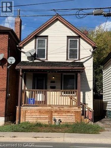 Two-story facade with light-colored siding, contrasting trim, a brick chimney, and a covered porch with wood railing - 1087 Cannon Street E, Hamilton, ON 