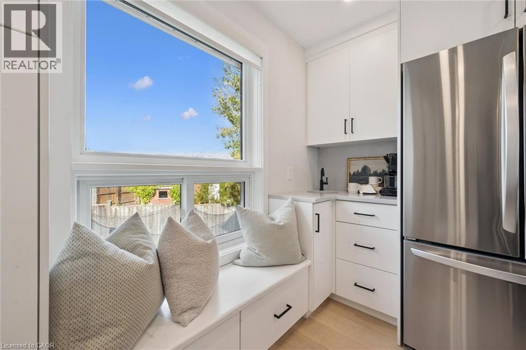 Built-in window bench with integrated storage, white cabinetry, stainless steel refrigerator, undermount sink with a matte black faucet, and light wood-finish flooring - 6 Branwood Place, Kitchener, ON