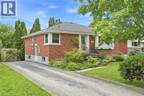 Red brick exterior featuring a hip roof, asphalt driveway, metal porch railings, and mature landscaping - 241 East 44Th Street, Hamilton, ON 