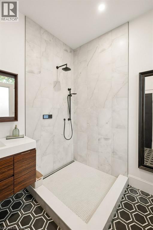 Walk-in shower featuring floor-to-ceiling large-format tile, matte black rainfall shower head with handheld attachment, custom stone-finish base with penny-round tiling, and floating wood-finish vanit - 361 Lee Circle, Waterloo, ON