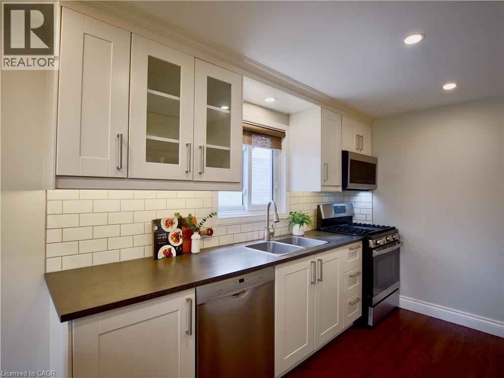 Kitchen featuring white shaker-style cabinetry with glass-panel inserts, subway tile backsplash, dark solid-surface countertops, stainless steel appliances, and wood-finish flooring - 449 Exmoor Street, Waterloo, ON