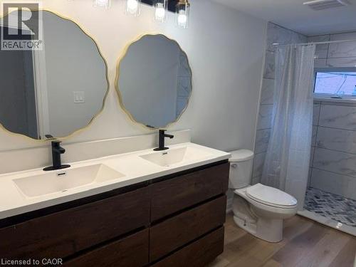 Double vanity featuring dark wood-finish cabinetry, a solid surface countertop, matte black fixtures, scalloped gold-rimmed mirrors, and a tiled shower enclosure with herringbone floor pattern - 197 Ferguson Avenue S, Hamilton, ON 