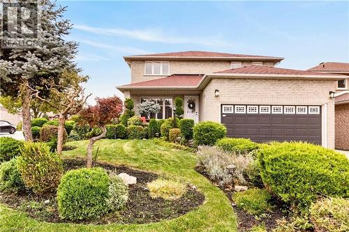 Two-story brick exterior featuring a multi-tonal masonry facade, a multi-car garage with decorative windows, a hip roof, and professional landscaping with mature trees and shaped shrubs - 53 Bastille Street, Hamilton, ON 