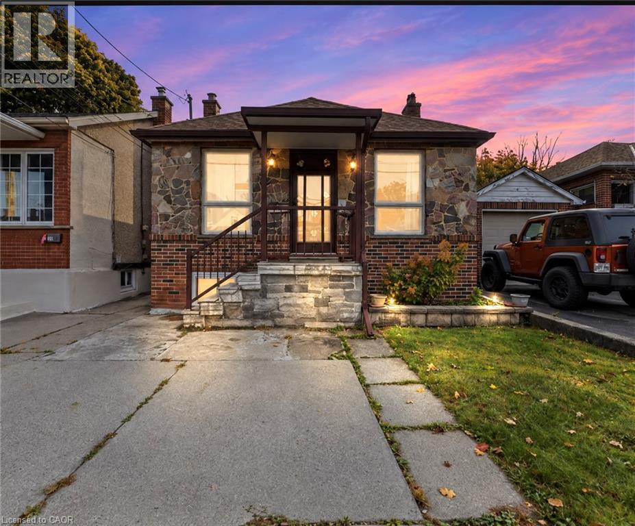 Exterior facade featuring stone and brick masonry, covered front porch with metal railings, concrete driveway, and attached garage - 81 East 23Rd Street, Hamilton, ON