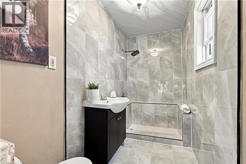 Full-height tiled walls featuring neutral-toned stone-finish tile, dark-finish wood cabinetry, integrated white porcelain sink, wall-mounted shower fixture, and plank-style ceiling detailing - 81 East 23Rd Street, Hamilton, ON 