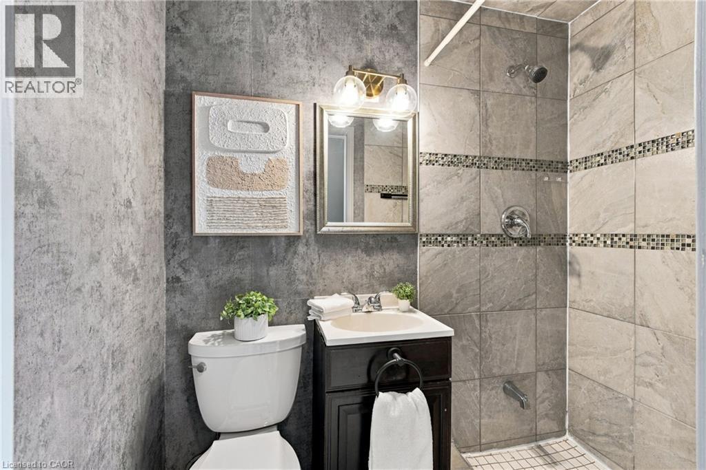 Bathroom featuring a dark wood-finish vanity, stone-patterned wall tile, glass mosaic tile accents, a metallic wall-mounted light fixture, and a built-in tub and shower combination - 81 East 23Rd Street, Hamilton, ON
