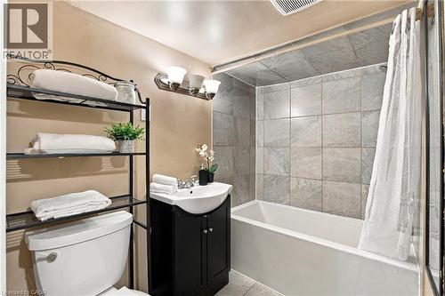 Bathroom featuring a vanity with a white basin and dark-finish cabinet, tub-shower combination with floor-to-ceiling stone-look tiling, and three-light wall-mounted fixture - 81 East 23Rd Street, Hamilton, ON 