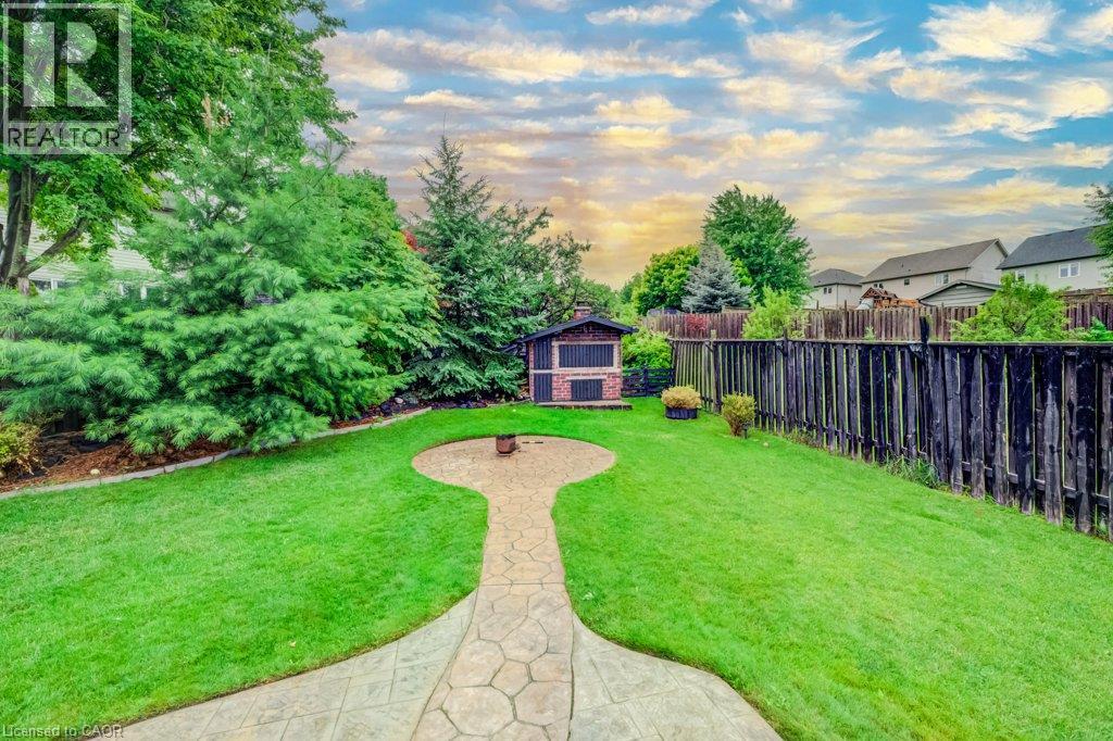 Backyard featuring a grass lawn, stamped concrete walkway, integrated circular fire pit area, detached storage structure, and mature evergreen trees enclosed by a wood privacy fence - 521 Snowdrop Court, Kitchener, ON