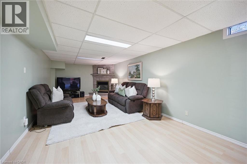 Neutral-toned room featuring a corner fireplace with stone surround, recessed drop ceiling, light-toned wood-finish flooring, and a single egress window - 11 Caledon Court, Cambridge, ON