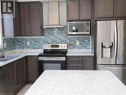 Kitchen featuring dark wood cabinetry, light-toned stone-finish countertops, multi-toned blue glass tile backsplash, stainless steel appliances, and a stainless steel wall-mounted range hood -