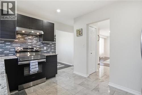 Kitchen featuring dark wood-finish cabinetry, stone-style countertops, multi-toned horizontal tile backsplash, stainless steel range with integrated hood, and polished tile flooring - 90 June Street, Hamilton, ON 