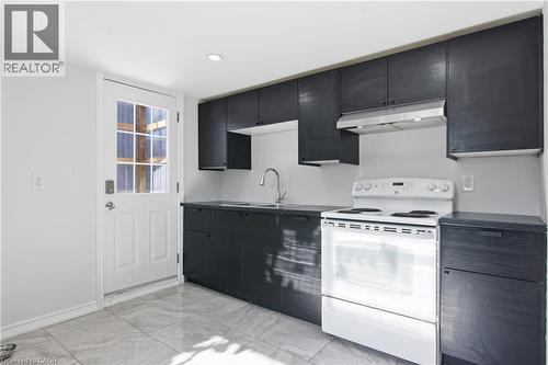 Kitchen featuring dark cabinetry, stone-finish countertops, tile flooring, a stainless steel faucet, and a white range with a range hood - 90 June Street, Hamilton, ON 
