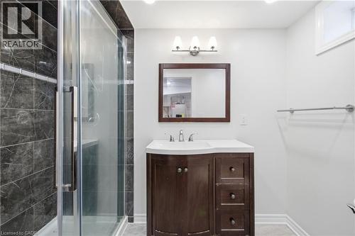 Bathroom featuring a dark wood-finish vanity with a white integrated sink, glass-enclosed shower with dark stone-finish wall tile, and a framed wall mirror with integrated three-bulb vanity light - 90 June Street, Hamilton, ON 