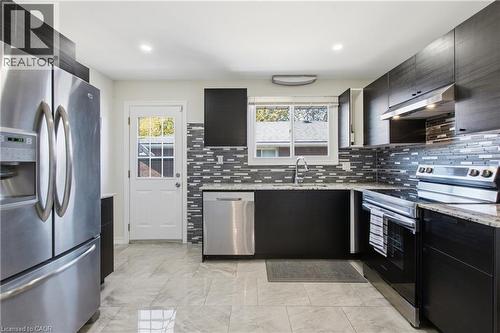 Kitchen featuring dark flat-panel cabinetry, stone-patterned countertops, stainless steel appliances, a multi-tonal linear tile backsplash, and ceramic tile flooring - 90 June Street, Hamilton, ON 