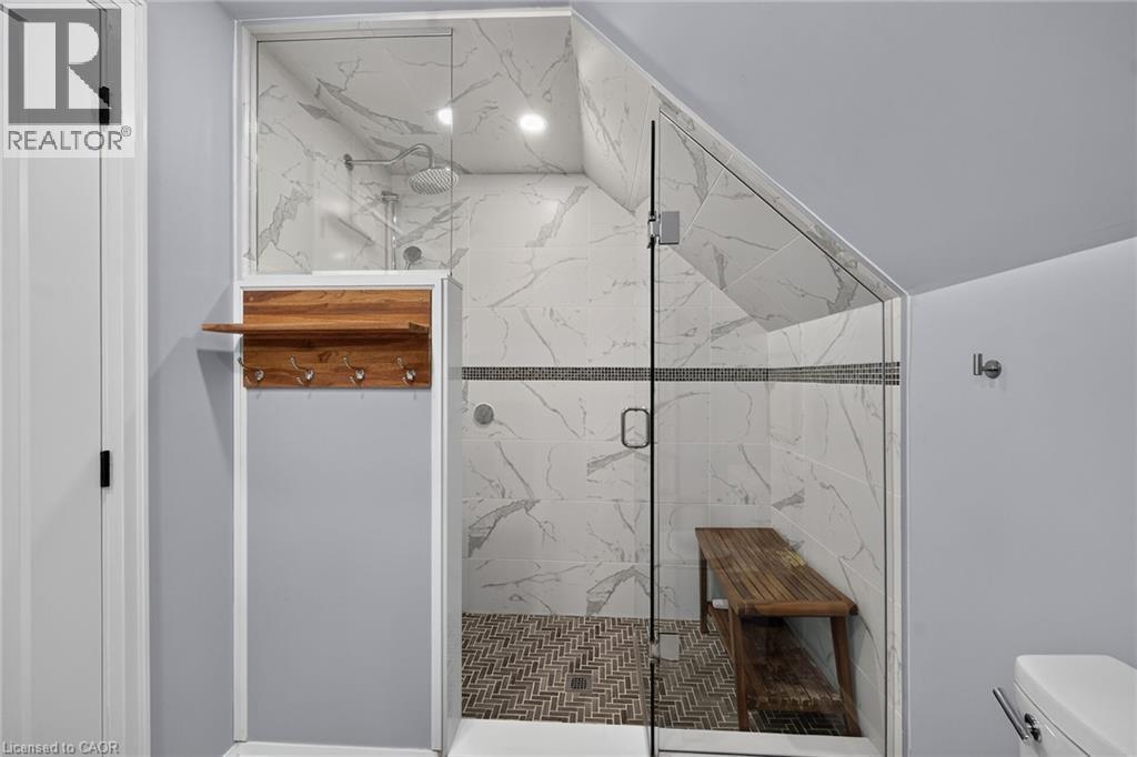 Walk-in shower featuring white veined tile walls, a herringbone-patterned tile floor, a glass enclosure, and a built-in recessed ceiling light. - 10 Mulock Avenue, Hamilton, ON
