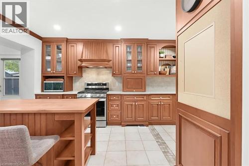 Wood-tone cabinetry with glass-front uppers, stainless steel range with overhead vent, light-toned backsplash, white countertops, and tile flooring with accent strip - 31 Berrisfield Crescent, Hamilton, ON 