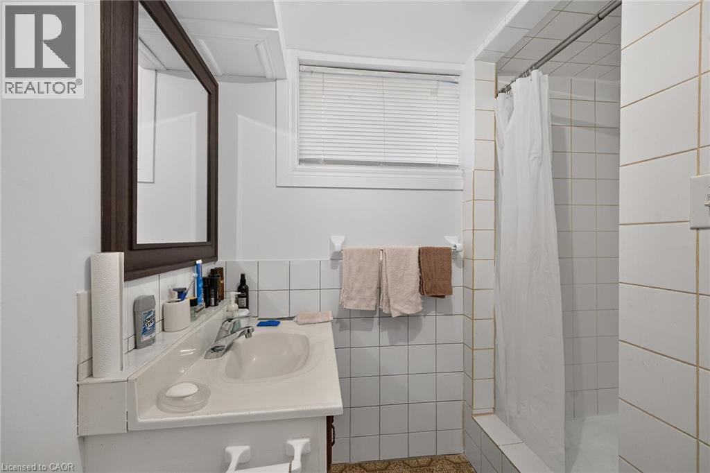 Bathroom featuring a vanity with an integrated sink, wall-mounted mirror, inset shower with white wall tiling, and a window with fitted blinds - 411 Queen Victoria Drive, Hamilton, ON
