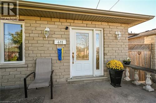 Exterior entrance featuring a light-colored masonry facade, a white entry door with an integrated glass sidelight, stone-finish porch, and dual wall-mounted coach lights - 411 Queen Victoria Drive, Hamilton, ON 