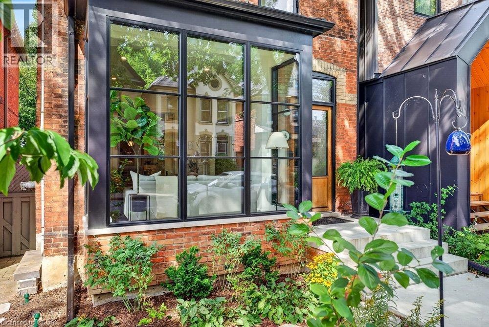 Brick exterior with black-framed large windows, stone steps, landscaped garden beds, and a wood-paneled entry door - 16 Bruce Street, Hamilton, ON