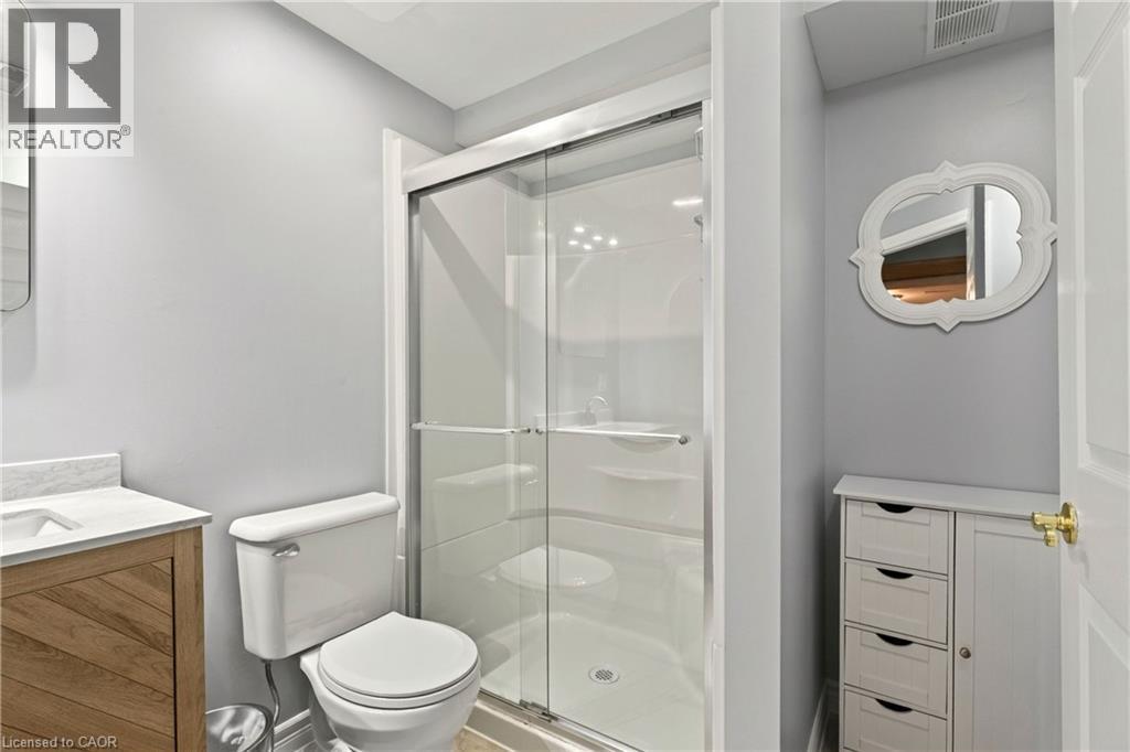 Glass-enclosed shower, white toilet, wood-finish vanity with countertop, white storage cabinet, and wall-mounted mirror - 31 Grindstone Way, Hamilton, ON