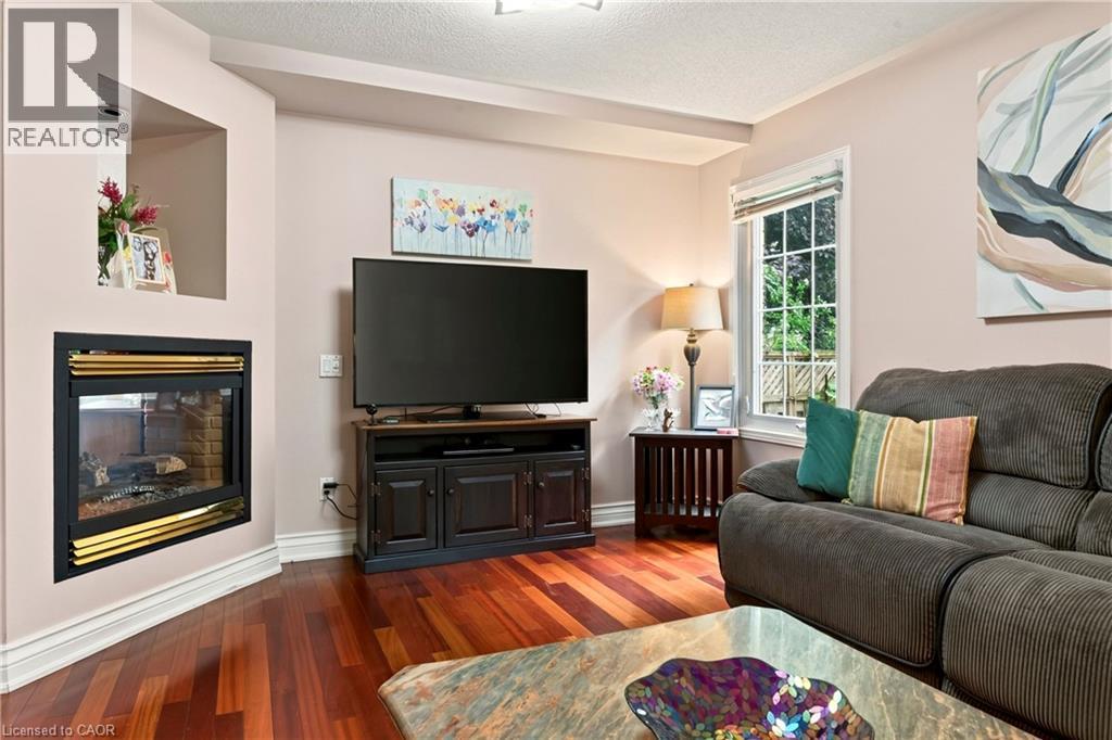 Corner fireplace with a brass-finish trim, wood-finish flooring, a window with a white frame, and light-toned walls - 31 Grindstone Way, Hamilton, ON