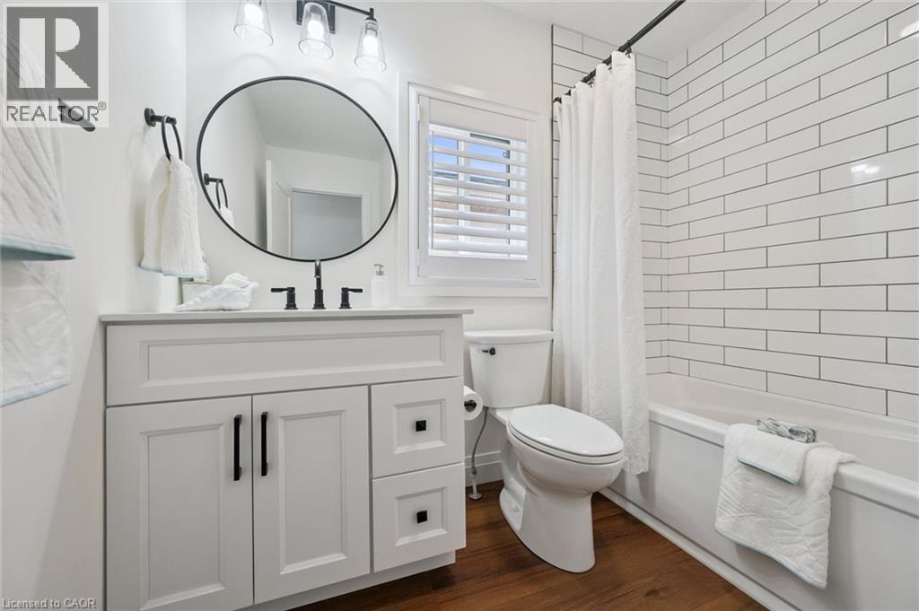 White vanity with black hardware, round mirror, dark wood-finish flooring, subway tile bath surround, and white plantation shutters - 2294 Seton Crescent, Burlington, ON