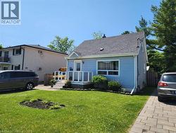 Light blue vinyl siding cottage featuring a covered front porch with white railings, peaked roofline with architectural detailing, manicured lawn with mature landscaping, paver driveway, and multi-car - 