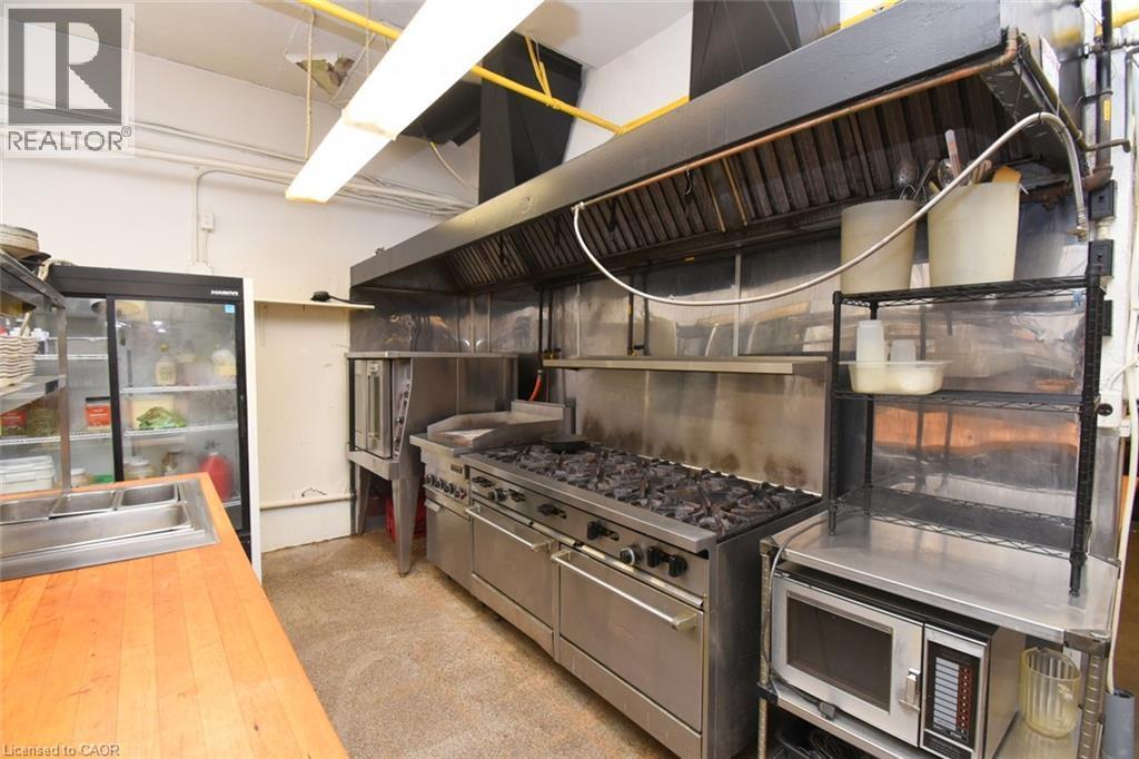 Professional-grade commercial kitchen featuring a six-burner range with double ovens, stainless steel backsplash and countertops, overhead exhaust hood system, and integrated refrigeration units - 942-946 Barton Street E, Hamilton, ON