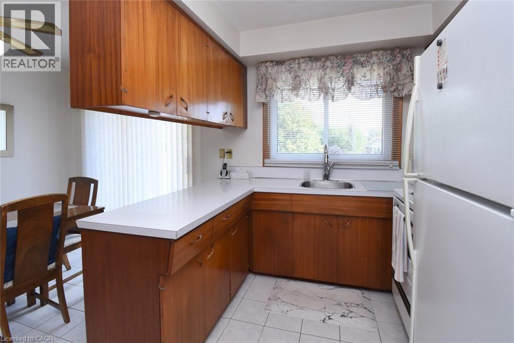 Compact kitchen featuring warm wood cabinetry, white laminate countertops, and an L-shaped layout with integrated sink and window overlooking natural light - 98 Kennedy Avenue, Hamilton, ON