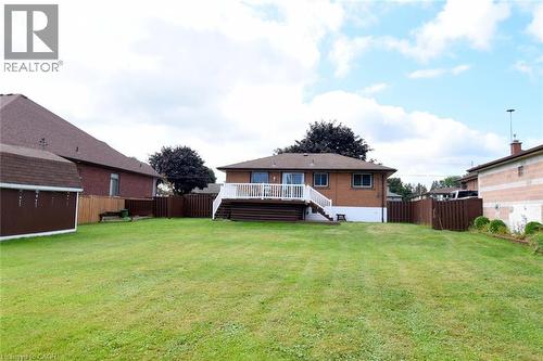 Expansive manicured lawn with mature landscaping trees, brick exterior residence featuring a covered front deck with railing, attached garage with wood-finish door, and wood-fenced perimeter - 98 Kennedy Avenue, Hamilton, ON 