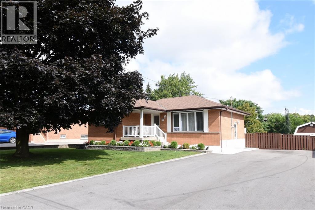 Brick exterior with a covered porch featuring white railings, mature front yard tree, paved driveway, and perimeter fencing - 98 Kennedy Avenue, Hamilton, ON