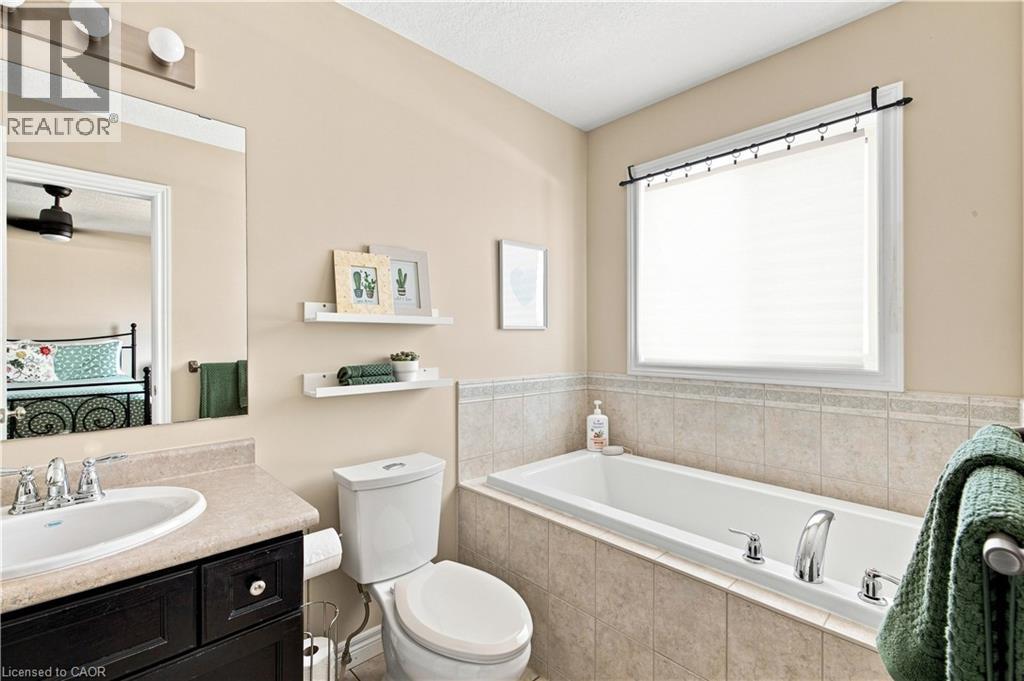 Built-in bathtub with tiled surround, single vanity with integrated sink, window with blinds, wall-mounted floating shelves, and toilet - 187 Eden Oak Trail, Kitchener, ON