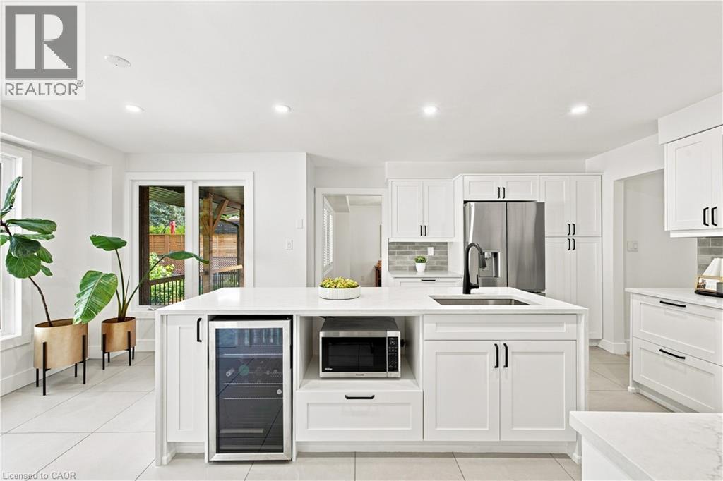White shaker-style cabinetry with matte black hardware, stainless steel appliances, a built-in wine refrigerator, a large center island with an undermount sink, and light-toned tile flooring - 62 Corfield Drive, Kitchener, ON
