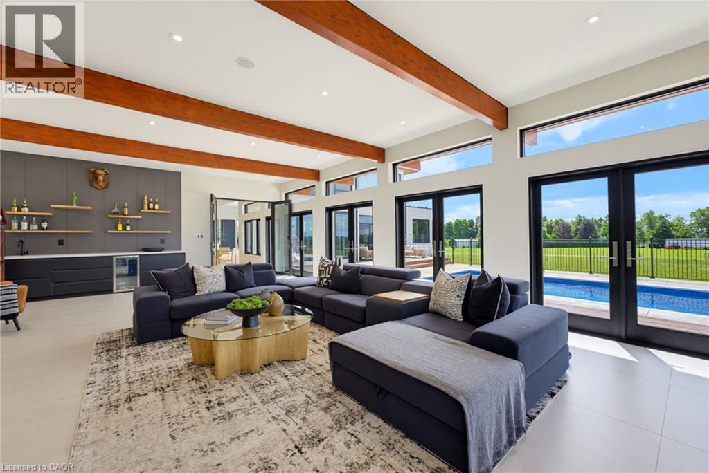 Sophisticated open-concept living space showcasing exposed wood beam ceiling, floor-to-ceiling black-framed glass doors and transom windows, polished concrete flooring, and a built-in wet bar with bra - 5709 Forth Line, Hillsburg, ON