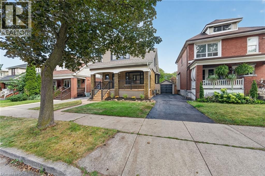 Brick exterior with white trim, covered front porch, dormer window, established front landscaping, and a paved driveway leading to a detached garage - 229 Park Row S, Hamilton, ON